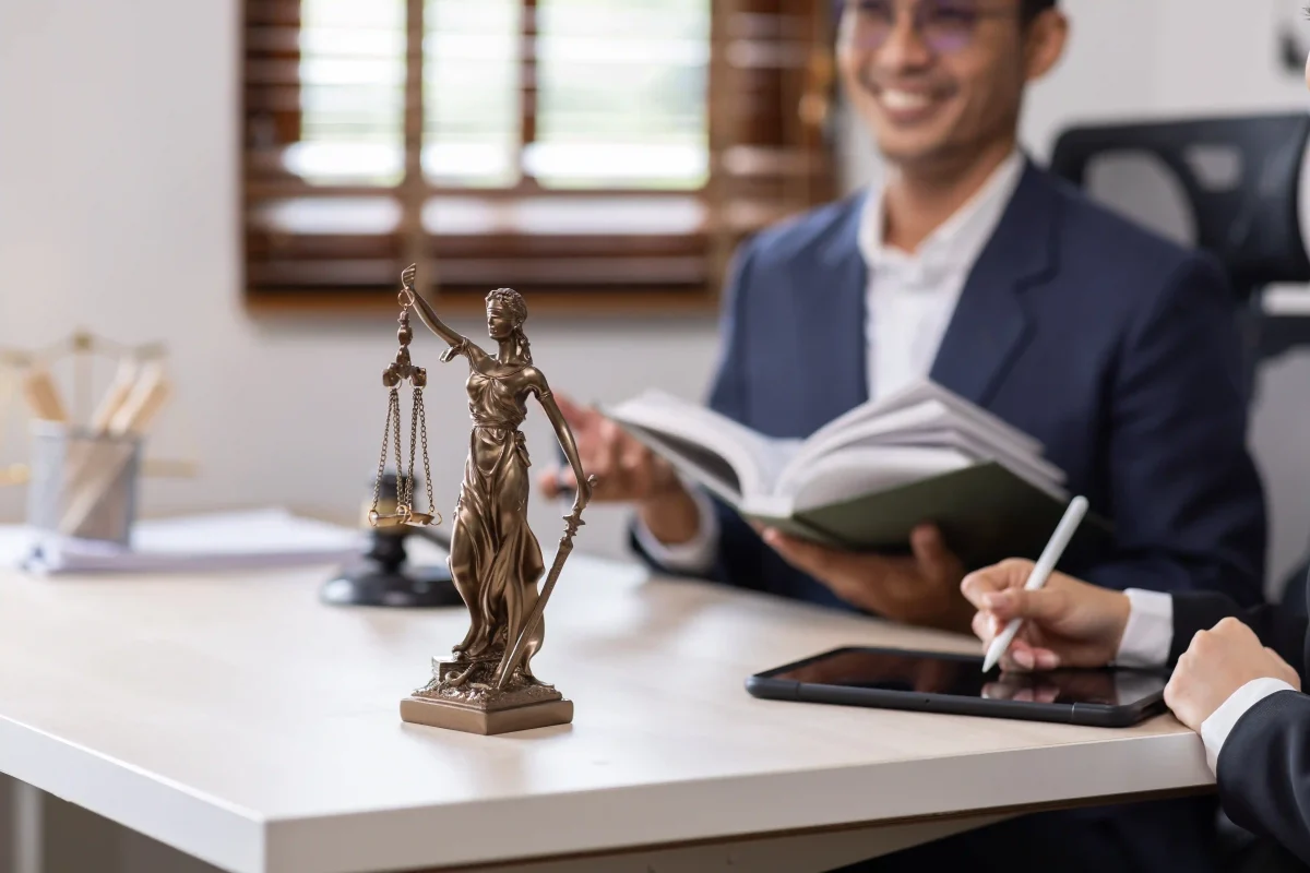 Estátua da deusa da justiça sobre mesa de escritório, com profissionais ao fundo analisando documentos — tema: anulação e revogação de licitação.